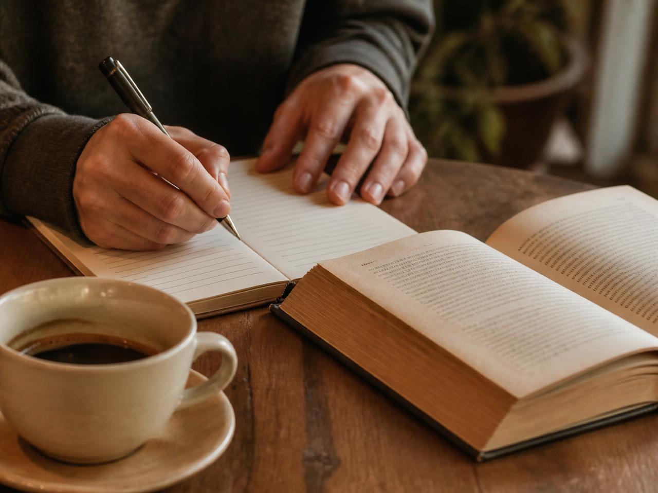 Mãos anotando à caneta em um caderno de papel ao lado de um livro aberto e uma xícara de café.